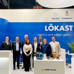 Auf dem Bild ist eine Gruppe von Personen in formeller Kleidung auf einem Messestand zu sehen, die freundlich in die Kamera lächeln. Im Hintergrund sieht man die Logos von Stannol und Lökast.