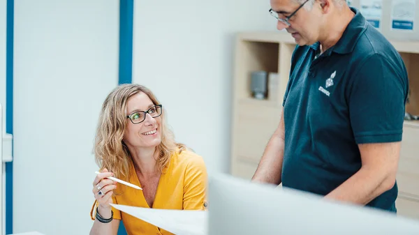 Eine Frau in Gelb und ein Mann in Blau diskutieren an einem Schreibtisch.