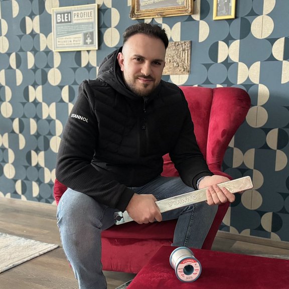 The picture shows a young, dark-haired man with a beard sitting on a red armchair and holding a plumb bob in his hand. He is wearing blue jeans and a black jacket.