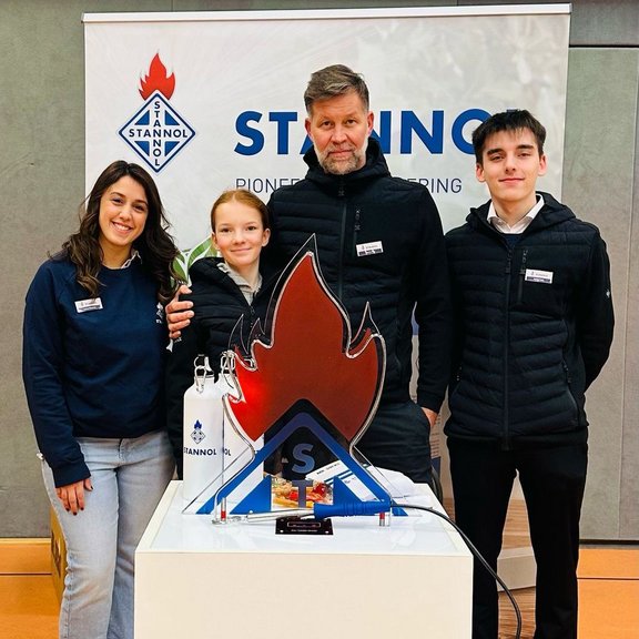 The picture shows four people in front of a Stannol banner, smiling friendly into the camera. In front of them is a table with a "hot wire" in the shape of the Stannol flame. 