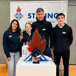 Auf dem Bild sind vier Personen vor einem Stannol-Banner zu sehen, die freundlich in die Kamera lächeln. Vor ihnen steht ein Tisch mit einem "heißen Draht" in Form der Stannol-Flamme. 