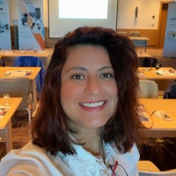 Auf dem Bild ist eine junge Frau mit braunen, schulterlangen Haaren in einer weißen Bluse zu sehen. Sie macht ein Selfie mit einem Konferenzraum im Hintergrund.