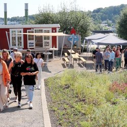 Auf dem Bild ist ein Garten mit einem Weg zu sehen, auf dem mehrere Personen spazieren gehen. Im Hintergrund ist ein roter Bauwagen zu sehen. 