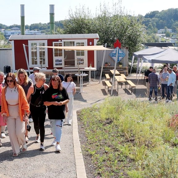 Auf dem Bild ist ein Garten mit einem Weg zu sehen, auf dem mehrere Personen spazieren gehen. Im Hintergrund ist ein roter Bauwagen zu sehen. 