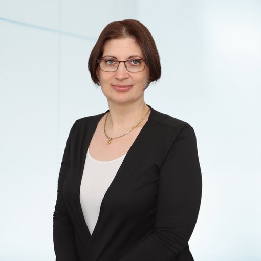 The picture shows a woman with a dark brown pageboy cut. She is wearing a white top and a black blazer. She is wearing a pair of glasses and a half-length gold chain around her neck. She smiles friendly into the camera. 