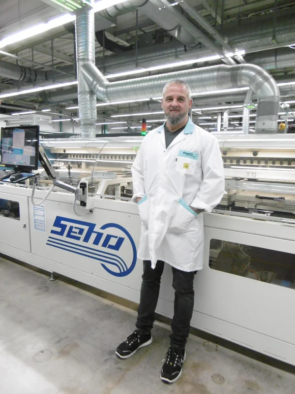 Auf dem Bid ist ein Mann mit grauen Haaren und Bart in einer Produktionshalle zu sehen. Er trägt einen weißen Kittel und lächelt freundlich in die Kamera.