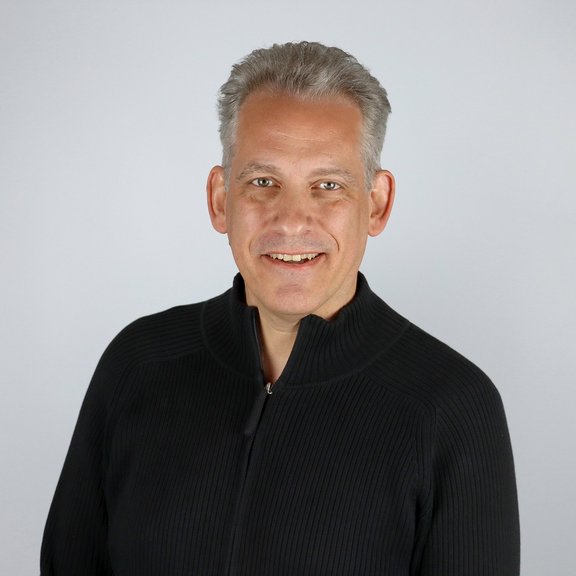 The picture shows a man with grey hair and a dark jumper. He smiles friendly into the camera.