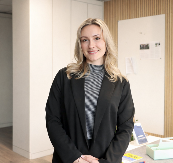 [Translate to Englisch:] Frau in schwarzem Blazer lächelt in modernem Büro.