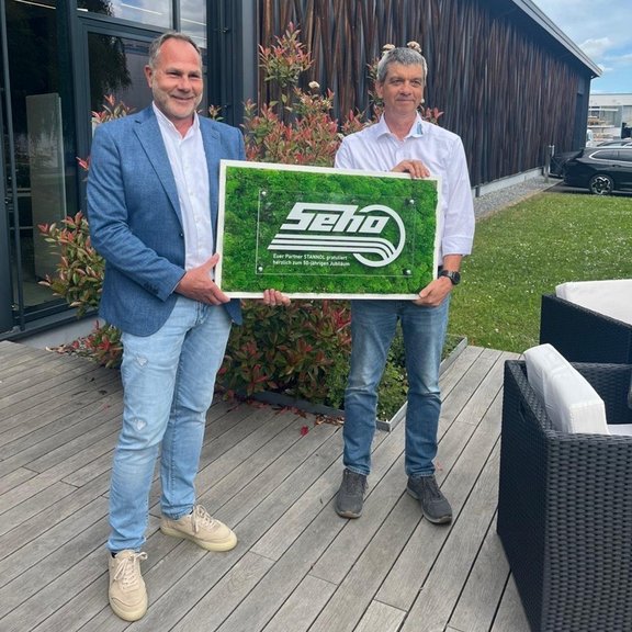 Zwei Männer halten ein Schild im Freien vor einem modernen Gebäude.