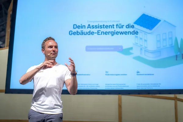 Auf dem Bild ist der Referent Pascal Biesenbach bei seinem Vortrag zu sehen. Er hat kurze blonde Haare und trägt ein weißes T-Shirt.