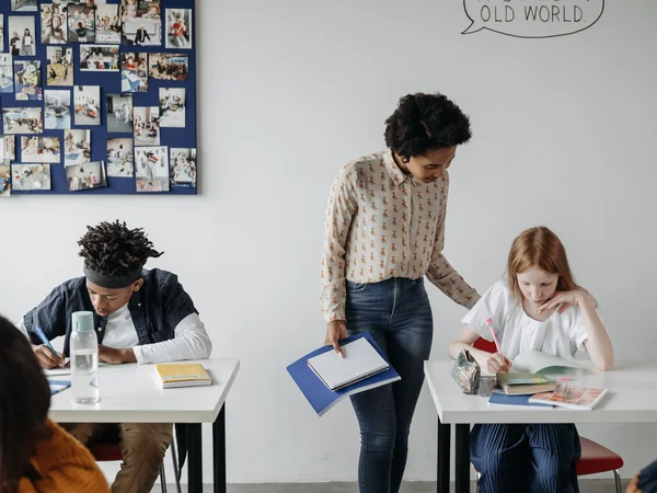 The image is a stock photo. A teacher is looking after two pupils who are each bent over a desk. 