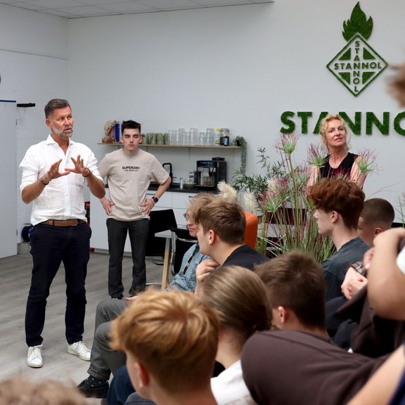 A group of young people sits attentively in a modern, bright room at Stannol. A man in a white shirt speaks engagingly in front of the group, while two other adults stand in the background and listen. The Stannol logo can be seen on the wall, with plants and a small coffee bar next to it. 