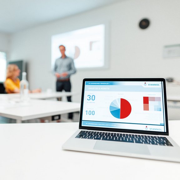 [Translate to Englisch:] Auf dem Bild ist ein Seminarraum zu sehen, im Vordergrund steht ein aufgeklappter Laptop, im Hintergrund sitzen zwei Seminar-Teilnehmerinnen und der Seminarleiter erklärt etwas vor einer Leinwand.