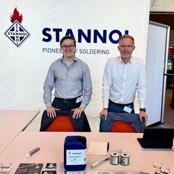 Auf dem Bild sind zwei Männer hinter einem Tisch zu sehen, die freundlich in die Kamera lächeln. Auf dem Tisch sind Produkte von Stannol wie ein Kanister Flussmittel, ein Lotbarren und Lötdraht-Spulen zu erkennen. Im Hintergund ist das Stannol-Logo zu sehen. 