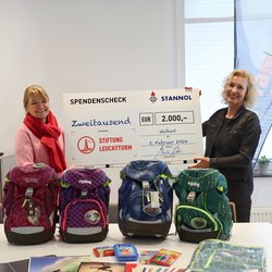 Auf dem Bild sind zwei Frauen zu sehen, die hinter einem Tisch stehen und gemeinsam einen Scheck in die Höhe halten. Auf dem Tisch stehen vier Schulranzen und weiteres Schulmaterial.