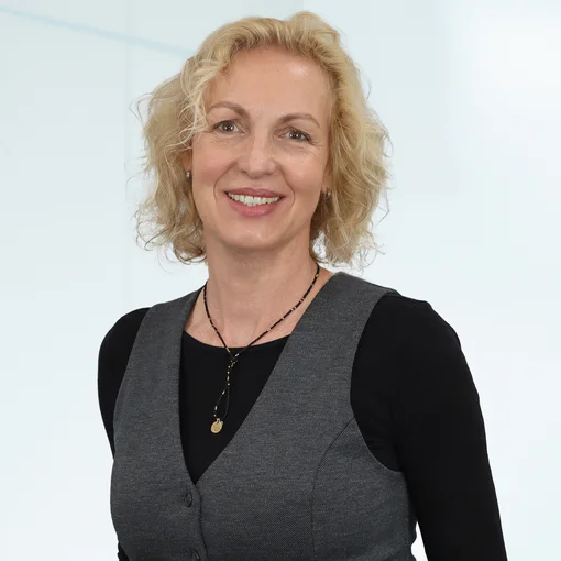 Auf dem Bild ist eine Frau mit hellblonden, halblangen welligen Haaren zu sehen. Sie trägt ein schwarzes Shirt, eine graue Weste und eine schwarz-goldene Kette um den Hals. Sie lächelt freundlich in die Kamera.