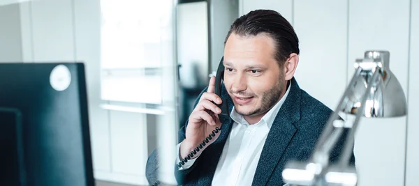 The picture shows a man sitting at a desk in front of a computer in an office, talking on the phone. He has brown hair, a beard and is wearing a white shirt and a dark-coloured suit jacket.