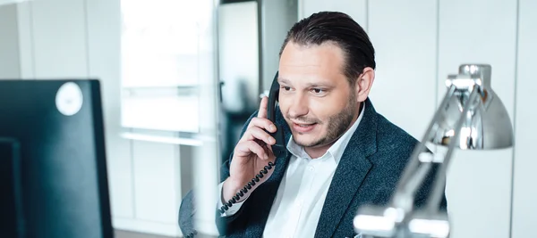 Auf dem Bild ist ein Mann zu sehen, der im Büro an einem Schreibtisch vor einem Computer sitzt und dabei telefoniert. Er hat braune Haare, einen Bart, trägt ein weißes Hemnd und ein dunkles Sakko.