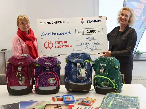 Das Bild zeigt Susanne Schlüter, Assistenz der Geschäftsführung von Stannol, wie sie Schulmaterial und einen Scheck an eine Mitarbeiterin der Stiftung Leuchtturm übergibt. Sie steht rechts neben einem Tisch, auf dem Schulranzen und andere Schulmaterialien abgestellt sind. Susanne Schlüter hat hellblonde Haare und trägt ein schwarzes Oberteil. Die Mitarbeiterin von Leuchtturm steht auf der linken Seite des Tisches und trägt ein rotes Oberteil und eine Brille. Beide Frauen lächeln freundlich in die Kamera. 
