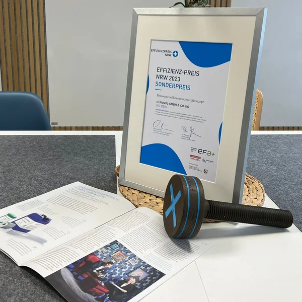 Auf dem Bild ist ein Tisch zu sehen, auf dem eine gerahmte Urkunde, ein aufgeschlagener Magazin-Artikel und eine Preis-Trophäe in Form einer großen hölzernen Schraube zu sehen ist.