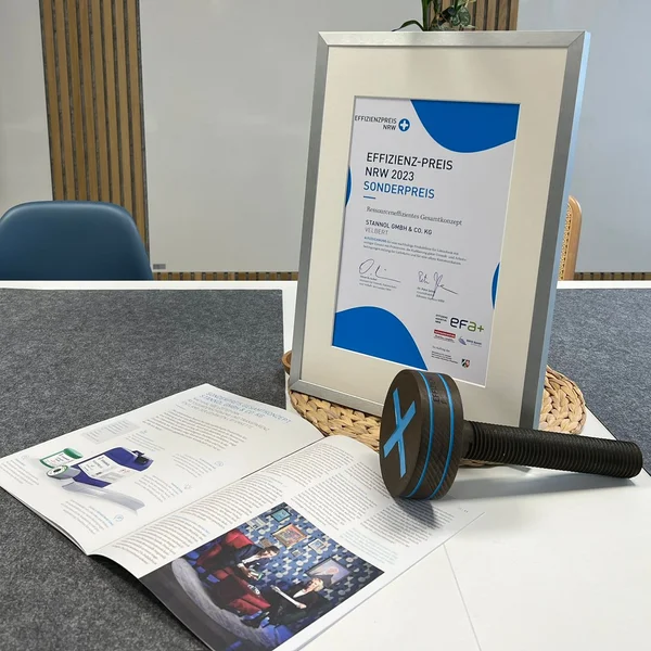 Auf dem Bild ist ein Tisch zu sehen, auf dem eine gerahmte Urkunde, ein aufgeschlagener Magazin-Artikel und eine Preis-Trophäe in Form einer großen hölzernen Schraube zu sehen ist.