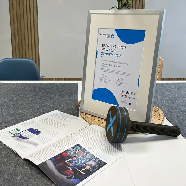 The picture shows a table on which a framed certificate, an open magazine article and a prize trophy in the form of a large wooden screw can be seen.