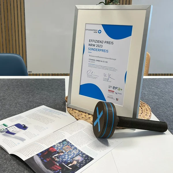 The picture shows a table on which a framed certificate, an open magazine article and a prize trophy in the form of a large wooden screw can be seen.