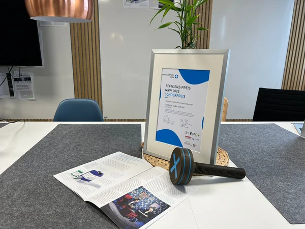 The picture shows a table on which a framed certificate, an open magazine article and a prize trophy in the form of a large wooden screw can be seen.