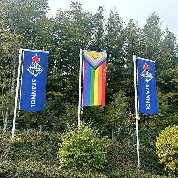 Auf dem Bild sind drei Flaggen zu sehen, rechts und links zwei blaue Stannol-Flaggen mit Logo und in der Mitte eine Regenbogenflagge.
