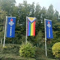 Auf dem Bild sind drei Flaggen zu sehen, rechts und links zwei blaue Stannol-Flaggen mit Logo und in der Mitte eine Regenbogenflagge.