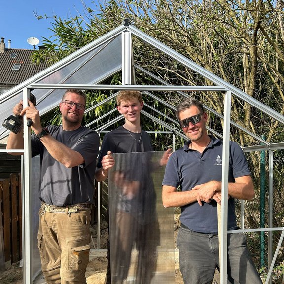 Auf dem Bild sind drei Männer in Arbeitskleidung zu sehen, die gemeinsam ein kleines Gewächshaus aufbauen. Sie lächeln freundlich in die Kamera.
