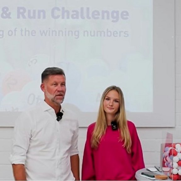 The picture shows a man and a young blonde woman. They are standing in front of a screen. Next to them is a table with a container full of small balls. 