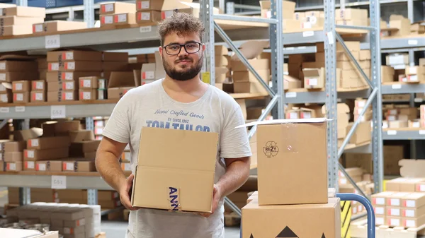 Mann in Lagerhalle, hält Karton, umgeben von Regalen mit weiteren Boxen.