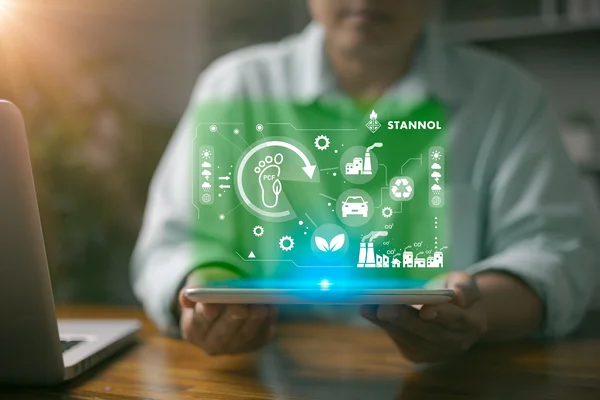 Das Bild zeigt eine Person, die ein Tablet in den Händen hält. Über dem Tablet schwebt eine grün leuchtende, digitale Darstellung, die verschiedene Symbole enthält, die für Nachhaltigkeit, Umweltschutz und industrielle Prozesse stehen. Dazu gehören unter anderem ein Fußabdruck mit dem Schriftzug PCF (Product Carbon Footprint), ein Recycling-Symbol, ein Blatt für Nachhaltigkeit sowie Symbole für Fabriken, Autos und Zahnräder. Oben rechts ist das Logo des Unternehmens Stannol zu sehen, das für innovative Lösungen in der Löttechnologie bekannt ist. Das Bild vermittelt den Fokus auf umweltfreundliche Prozesse, nachhaltige Innovationen und technologische Entwicklung. Die grüne Farbgebung unterstreicht den ökologischen und zukunftsorientierten Aspekt.