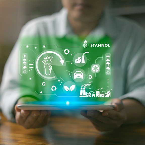 The image shows a person holding a tablet in their hands. Hovering above the tablet is a glowing green digital representation containing various symbols that symbolise sustainability, environmental protection and industrial processes. These include a footprint with the words PCF (Product Carbon Footprint), a recycling symbol, a leaf for sustainability and symbols for factories, cars and gears. At the top right is the logo of Stannol, a company known for innovative solutions in soldering technology. The image conveys the focus on environmentally friendly processes, sustainable innovation and technological development. The green colour scheme underlines the ecological and future-oriented aspect. Translated with DeepL.com (free version)