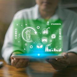 Das Bild zeigt eine Person, die ein Tablet in den Händen hält. Über dem Tablet schwebt eine grün leuchtende, digitale Darstellung, die verschiedene Symbole enthält, die für Nachhaltigkeit, Umweltschutz und industrielle Prozesse stehen. Dazu gehören unter anderem ein Fußabdruck mit dem Schriftzug PCF (Product Carbon Footprint), ein Recycling-Symbol, ein Blatt für Nachhaltigkeit sowie Symbole für Fabriken, Autos und Zahnräder. Oben rechts ist das Logo des Unternehmens Stannol zu sehen, das für innovative Lösungen in der Löttechnologie bekannt ist. Das Bild vermittelt den Fokus auf umweltfreundliche Prozesse, nachhaltige Innovationen und technologische Entwicklung. Die grüne Farbgebung unterstreicht den ökologischen und zukunftsorientierten Aspekt.