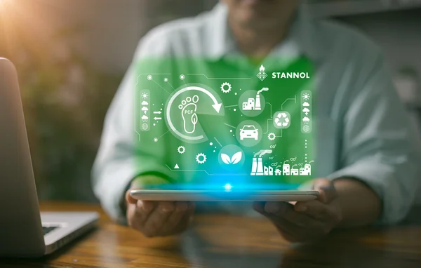 The image shows a person holding a tablet in their hands. Hovering above the tablet is a glowing green digital representation containing various symbols that symbolise sustainability, environmental protection and industrial processes. These include a footprint with the words PCF (Product Carbon Footprint), a recycling symbol, a leaf for sustainability and symbols for factories, cars and gears. At the top right is the logo of Stannol, a company known for innovative solutions in soldering technology. The image conveys the focus on environmentally friendly processes, sustainable innovation and technological development. The green colour scheme underlines the ecological and future-oriented aspect.