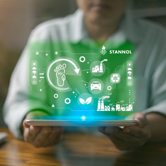 The image shows a person holding a tablet in their hands. Hovering above the tablet is a glowing green digital representation containing various symbols that symbolise sustainability, environmental protection and industrial processes. These include a footprint with the words PCF (Product Carbon Footprint), a recycling symbol, a leaf for sustainability and symbols for factories, cars and gears. At the top right is the logo of Stannol, a company known for innovative solutions in soldering technology. The image conveys the focus on environmentally friendly processes, sustainable innovation and technological development. The green colour scheme underlines the ecological and future-oriented aspect.