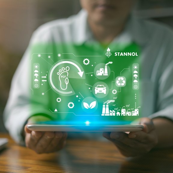 Das Bild zeigt eine Person, die ein Tablet in den Händen hält. Über dem Tablet schwebt eine grün leuchtende, digitale Darstellung, die verschiedene Symbole enthält, die für Nachhaltigkeit, Umweltschutz und industrielle Prozesse stehen. Dazu gehören unter anderem ein Fußabdruck mit dem Schriftzug PCF (Product Carbon Footprint), ein Recycling-Symbol, ein Blatt für Nachhaltigkeit sowie Symbole für Fabriken, Autos und Zahnräder. Oben rechts ist das Logo des Unternehmens Stannol zu sehen, das für innovative Lösungen in der Löttechnologie bekannt ist. Das Bild vermittelt den Fokus auf umweltfreundliche Prozesse, nachhaltige Innovationen und technologische Entwicklung. Die grüne Farbgebung unterstreicht den ökologischen und zukunftsorientierten Aspekt.