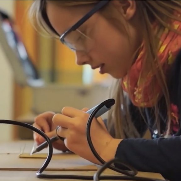 The picture shows a girl with brown hair and protective goggles holding a soldering iron. She is focussed on her work.
