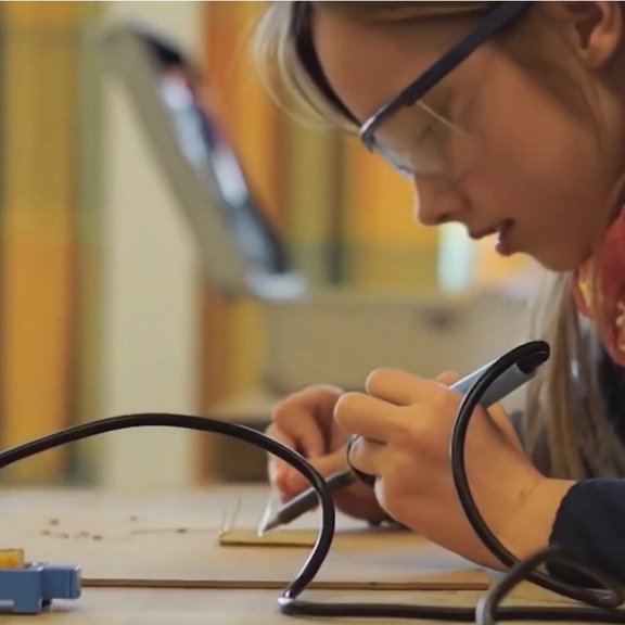 Auf dem Bild ist ein Mädchen mit braunen Haaren und einer Scutzbrille zu sehen, die einen Lötkolben in der Hand hält. Sie schaut konzentriert auf ihre Arbeit.
