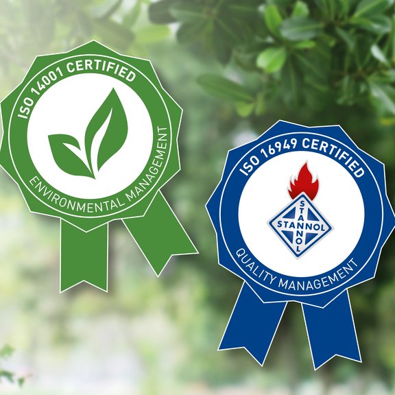 The image shows two certification badges side by side, placed against a blurred green background with leaves. On the left, there is a green badge in the shape of a rosette with the text "ISO 14001 Certified" at the top and "Environmental Management" at the bottom. In the center of the badge, there is a leaf symbol representing environmental friendliness. On the right, there is a blue badge in a similar rosette shape. It features the text "ISO 16949 Certified" at the top and "Quality Management" at the bottom. In the center of the badge, there is the logo of a company (Stannol) that includes a red flame symbol and a diamond shape. Both badges symbolize international certifications for environmental management (green) and quality management (blue).