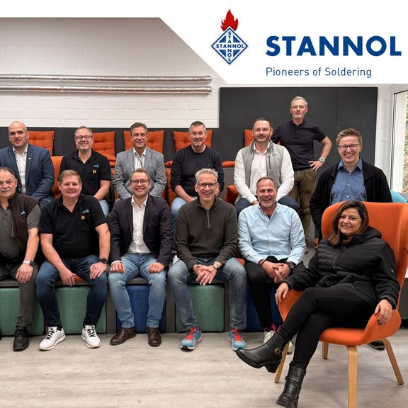 Gruppe von Personen in einem Raum, lächelnd, vor dem Stannol-Logo und Pflanzen.