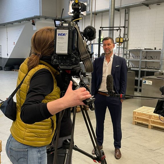 The picture shows a camerawoman filming Managing Director Marco Dörr for a television programme. They are standing in the Stannol production hall.