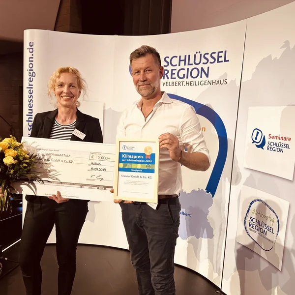 The picture shows a man and a woman holding a large cheque, a certificate and a bouquet of flowers. They are standing in front of a display of the key region.