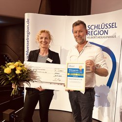Auf dem Bild sind Geschäftsführer Marco Dörr und die Assistentin der Geschäftsführung, Susanne Schlüter, zu sehen. Sie halten einen großen Scheck, einen Blumenstrauß und eine Urkunde in den Händen und lächeln freundlich in die Kamera. Im Hintergrund ist ein Aufsteller der Schlüsselregion zu erkennen.