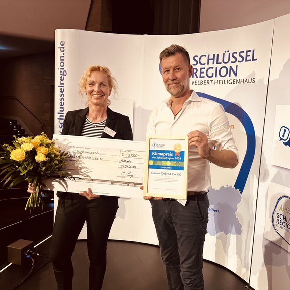 Auf dem Bild sind Geschäftsführer Marco Dörr und die Assistentin der Geschäftsführung, Susanne Schlüter, zu sehen. Sie halten einen großen Scheck, einen Blumenstrauß und eine Urkunde in den Händen und lächeln freundlich in die Kamera. Im Hintergrund ist ein Aufsteller der Schlüsselregion zu erkennen.