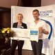 The picture shows Managing Director Marco Dörr and Management Assistant Susanne Schlüter. They are holding a large cheque, a bouquet of flowers and a certificate in their hands and smiling friendly into the camera. A display of the key region can be seen in the background.