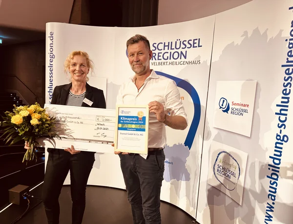 The picture shows a man and a woman holding a large cheque, a certificate and a bouquet of flowers. They are standing in front of a display of the key region.