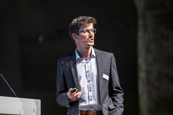 Auf dem Bild ist Quentin Zapf bei seienm Vortrag auf der Bühne zu sehen. Er hat braune Haare, trägt einen dunkelgrauen Anzug und ein helles Hemd.